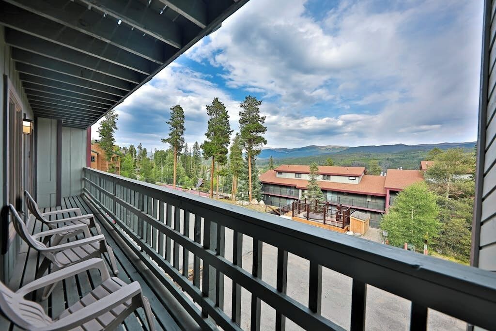 A balcony with a view of a mountainous landscape, a building with a red roof, and several trees.