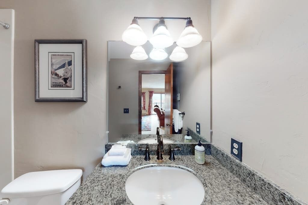 Une salle de bain avec des toilettes blanches, un lavabo sur un plan de travail en granit, un miroir avec éclairage intégré et un tableau encadré au mur.