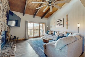 Una sala de estar con pared de piedra, ventilador de techo, sofá, mesa de centro y un cartel que dice 'PRAY FOR SNOW'.