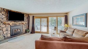 A living room with a stone fireplace, a flat-screen TV, a sofa, and a sliding glass door leading to an outdoor patio. - Rustic mountain escape with fireplace, W/D, hot tub, & grill - near skiing (Breckenridge)