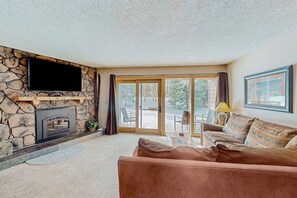 A living room with a stone fireplace, a flat-screen TV, a sofa, and a sliding glass door leading to an outdoor patio. - Rustic mountain escape with fireplace, W/D, hot tub, & grill - near skiing (Breckenridge)
