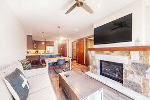 Ein modernes Wohnzimmer mit Kamin, einem flachbildfernseher darüber, einem Essbereich mit Tisch und Stühlen sowie einer Küche im Hintergrund.