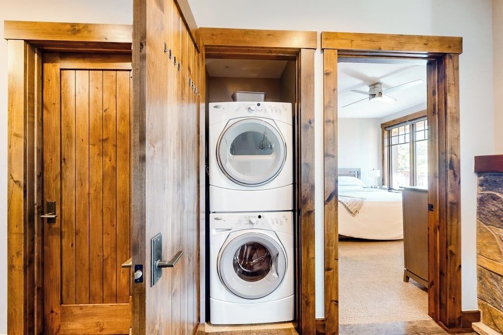 A laundry room with two stacked washers and dryers, wooden doors, and a view into a bedroom with a bed and a window.