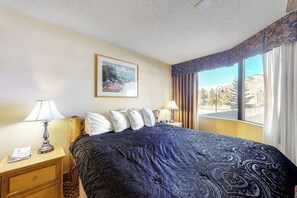Ein Hotelzimmer mit einem Bett, zwei Nachttischen, einer Lampe, einem gerahmten Bild an der Wand und einem Fenster mit Blick auf Bäume und Berge.