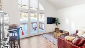 A living room with a wooden floor, a brown leather sofa, a TV mounted on the wall, and a sliding glass door leading to a deck.