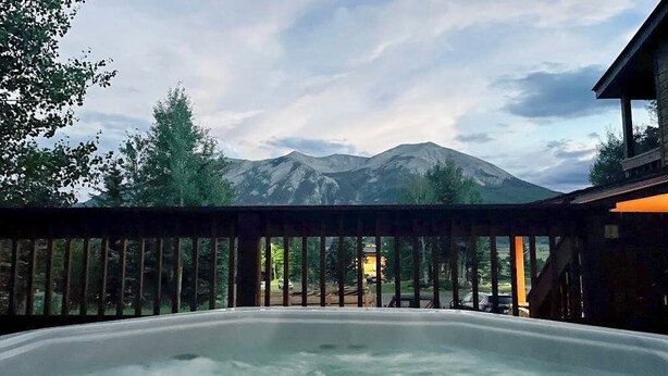 A hot tub with a mountain view, a wooden deck, and a building in the background.