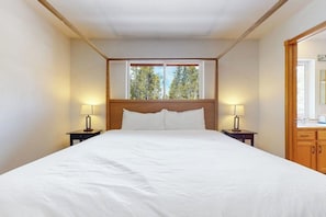 A bedroom with a large bed, two bedside lamps, a window with a view of trees, and wooden headboard and cabinetry. - Dog-friendly home with large deck, hot tub, woodstove, W/D, & serene setting (Fairplay)
