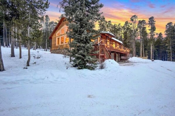 A log cabin with a deck, surrounded by snow and trees. - Dog-friendly home with large deck, hot tub, woodstove, W/D, & serene setting (Fairplay)