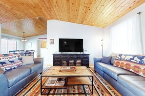 A living room with a wooden ceiling, a large flat-screen TV, blue sofas, a coffee table, and patterned rugs.