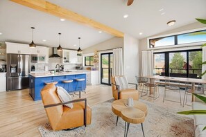 A modern kitchen with a central island, bar stools, and a dining area with a large window. - Modern mountain retreat with decks, kitchens, & kid-friendly items - near skiing (Crested Butte)