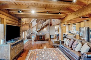 Ein Holzblockhaus-Interieur mit einer Holztreppe, einem Wohnzimmer mit Ledersofa, einem Flachbildfernseher und einer Küchenzeile.