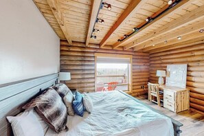 Ein Schlafzimmer in einer Blockhütte mit einem Bett, Kissen, einem Schreibtisch und einem Fenster mit Blick auf einen Balkon.
