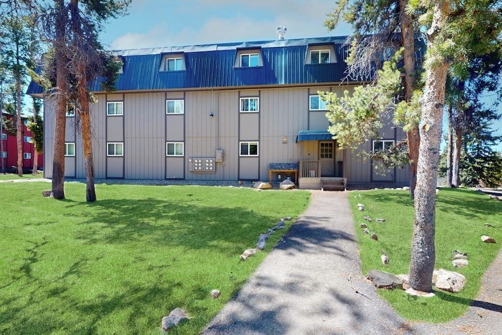 Un edificio de varios pisos con techo azul, entrada pavimentada y césped cuidado con árboles.