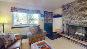 Una sala de estar con chimenea de piedra, un sillón estampado, un sofá y una ventana con vistas a árboles.