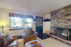 A living room with a stone fireplace, a patterned armchair, a sofa, and a window with a view of trees. - Quaint getaway steps from Main Street with fireplace & full kitchen (Winter Park)