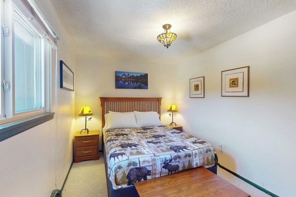 A bedroom with a bed, bedside lamps, a nightstand, a wooden headboard, a picture on the wall, and a window.