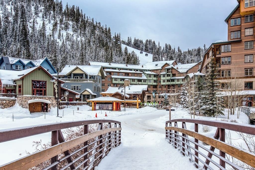 A snow-covered lodge with multiple buildings, a wooden bridge, and a mountainous backdrop.