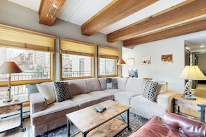 Ein Wohnzimmer mit einem Ecksofa, einem Couchtisch und einem roten Lederhocker. Zwei Fenster mit Jalousien, eine Lampe auf einem Beistelltisch und ein an der Wand befestigtes Fernsehgerät.
