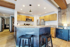 Eine moderne Küche mit Holzausbau, einer zentralen Kochinsel mit Hockern, einer Mikrowelle, einem Kühlschrank und einer Pendelleuchte.