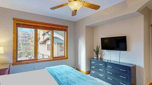 Quarto com cama, cômoda, televisão e janela com persianas.