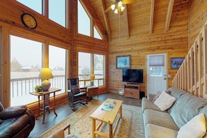 Interior de una cabaña de madera con sofá, mesa, televisor y ventanas amplias.
