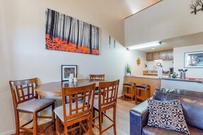 A dining area with a round wooden table and chairs, a kitchen in the background, and a living area with a leather couch.