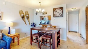 Een eetgedeelte met een houten tafel, vier krukken, een klok aan de muur en een vloerventilator.