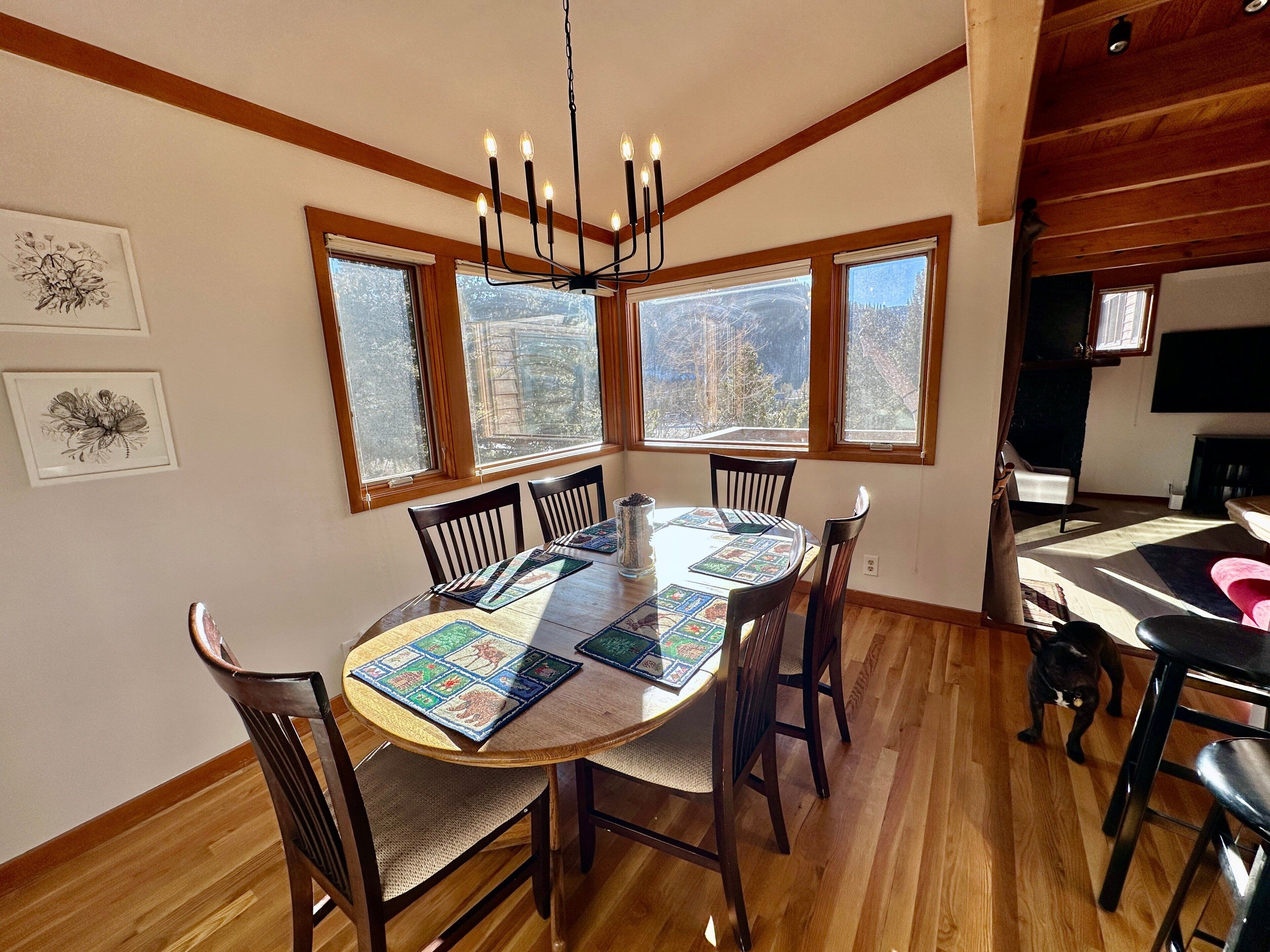 A dining area with a round table, chairs, a chandelier, and large windows with a view of trees.