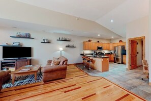 A modern living room with a sofa, TV, and coffee table, a kitchen with wooden cabinets and appliances, and a dining area with stools. - Convenient condo with fireplace & WiFi - walk to rec center pool, tennis & town (Pagosa Springs)