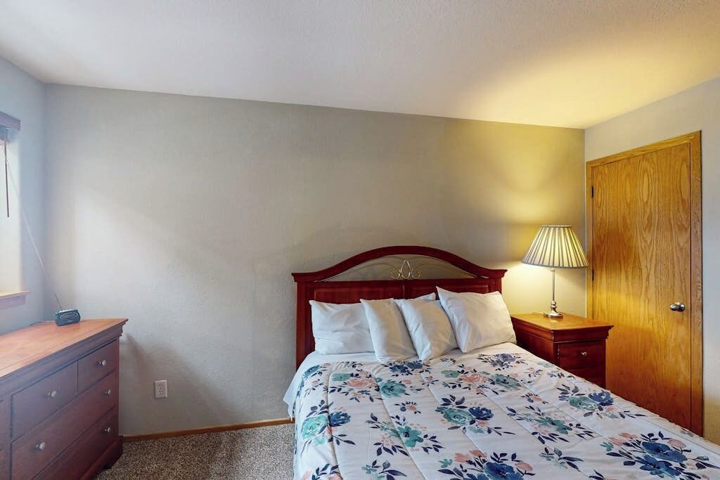 A bedroom with a wooden headboard, a floral bedspread, a bedside lamp, a wooden dresser, and a door.
