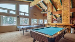Mesa de billar, dardos y zona de bar en una habitación con ventanas amplias y vigas de madera.