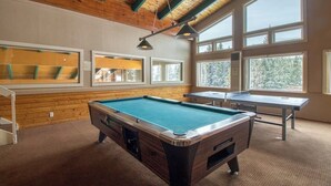 Una mesa de billar, una mesa de ping pong y una mesa de carambola en una sala con paredes de madera y ventanas amplias.