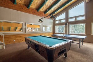 Ein Billardtisch, ein Tischtennis-Tisch und ein Snookertisch in einem Raum mit Holzwänden und großen Fenstern.