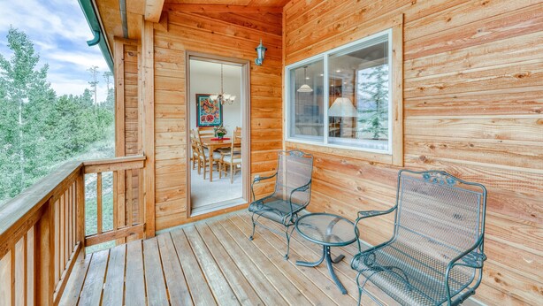 Une cabane en bois dotée d’un balcon, de deux chaises en métal, d’une table et d’une porte donnant sur un espace intérieur comprenant une table à manger et des chaises.