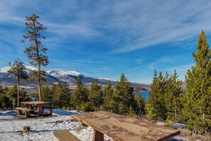Une table et un banc de pique-nique en bois, situés dans un paysage enneigé, avec des arbres et une montagne en arrière-plan.