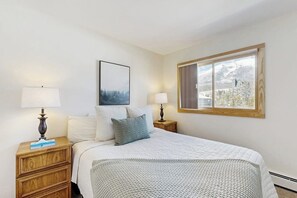A bedroom with a bed, two bedside tables, lamps, a framed picture, and a window with a view of snow-covered trees. - Spacious condo with shared pool & hot tub - great location (Silverthorne)