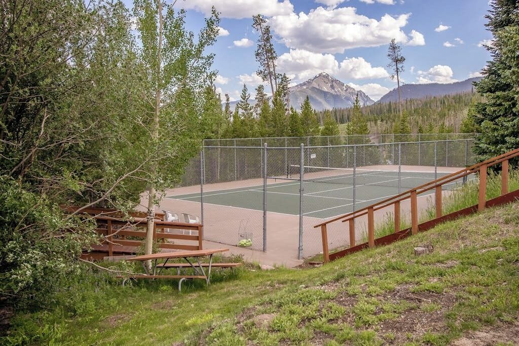 A tennis court with a fence, picnic tables, and a wooden bench.