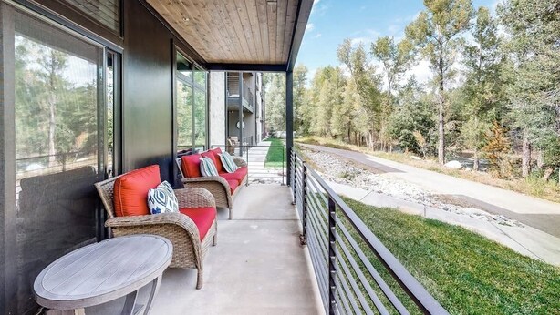A covered outdoor seating area with wicker furniture and a table, overlooking a road and trees. - Modern riverfront condo w/ a gas fireplace, washer/dryer & private patio (Silverthorne)