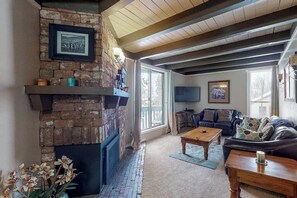 A living room with a brick fireplace, a couch, a coffee table, and a television. - Family-friendly condo w/ shared pool, hot tub, game room, & fitness room (Silverthorne)