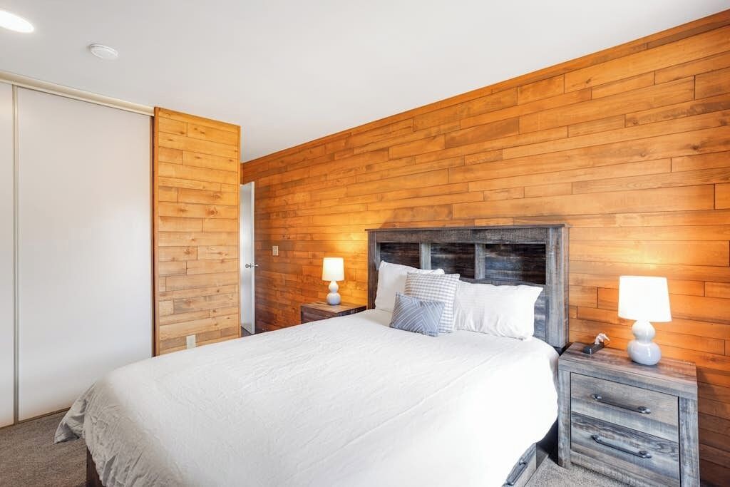 A bedroom with a wooden wall, a bed with white bedding, two bedside tables with lamps, and a wooden wardrobe.