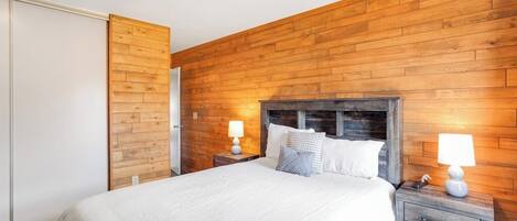 A bedroom with a wooden wall, a bed with white bedding, two bedside tables with lamps, and a wooden wardrobe.