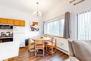 Eine moderne Küche mit Holzausstattung, einem Esstisch mit Stühlen, einem großen Fenster mit Vorhängen und einem Sofa in einem hellen Raum.