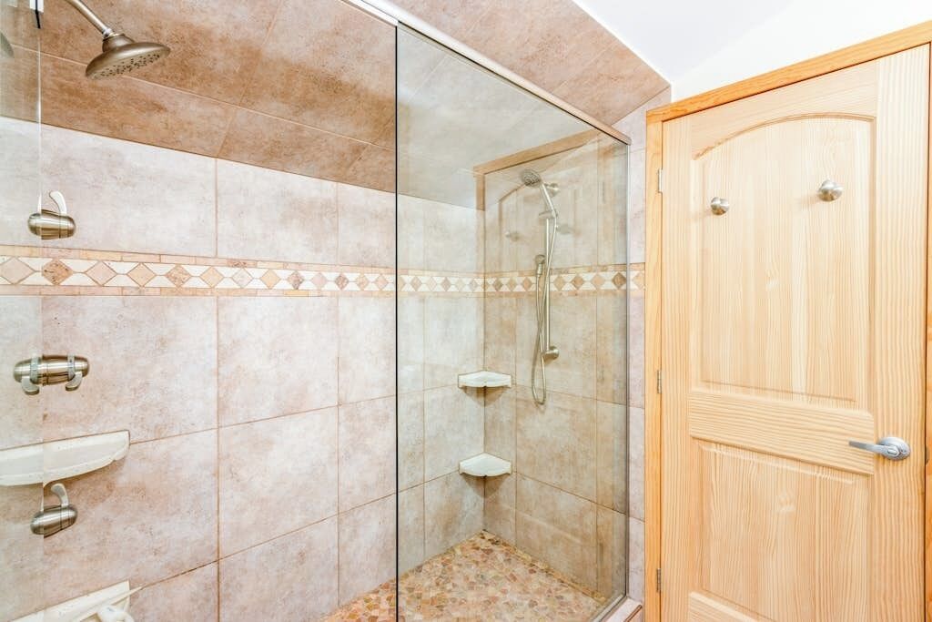 A shower with a glass door, a wooden door, and a tiled floor.