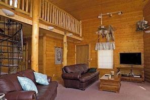 Ein Holzblockhaus-Interieur mit einem Wohnzimmer, das zwei Ledersofas, einen Couchtisch, einen Fernseher und einen Balkon mit Geländer zeigt.