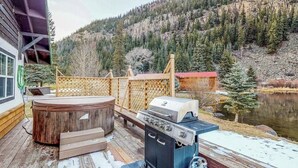 Une terrasse en bois dotée d’un jacuzzi, d’un barbecue et d’un espace salon extérieur, le tout offrant une vue sur un lac et des montagnes boisées.