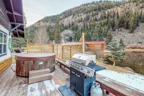 Eine Holzterrasse mit Whirlpool, Grill und Sitzgelegenheit im Freien mit Blick auf einen See und bewaldete Berge.