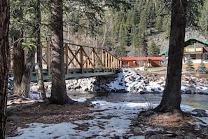 Eine Holzbrücke über einem Fluss, umgeben von Bäumen und einer Hütte mit rotem Dach.