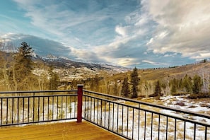 Eine Holzdar Terrasse mit Metallgeländer, die auf eine gebirgige Landschaft mit schneebedeckten Gipfeln und klarem Himmel blickt.