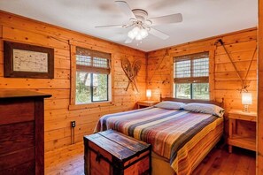Un dormitorio de cabaña de madera con una cama, dos ventanas, un ventilador de techo y un mapa enmarcado en la pared.