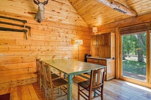 A wooden cabin interior with a dining table, chairs, a kitchen area, and a large window. - Dog-Friendly Cabin w/ Washer/Dryer, WiFi, & Firepit - Steps From the Rio Grande (South Fork)